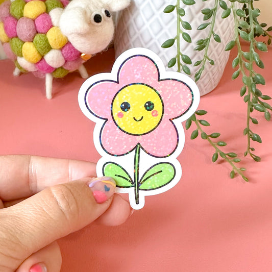 Hand holding a smiling flower sticker with a pink background featuring a sheep and plants.