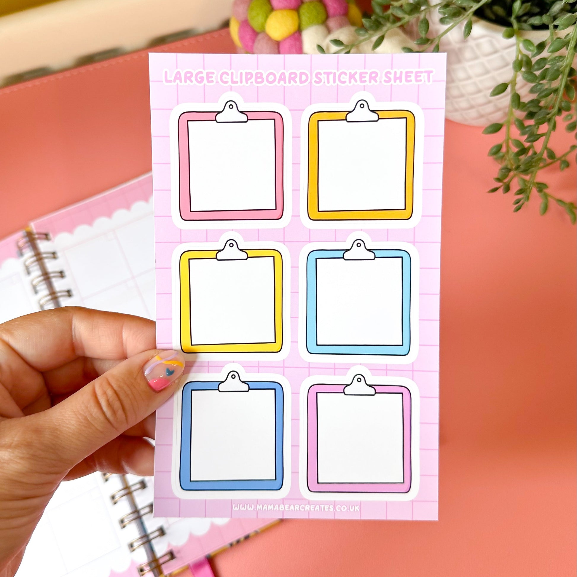 Sheet of large clipboard stickers held by a hand with a notebook and decorative items in the background.