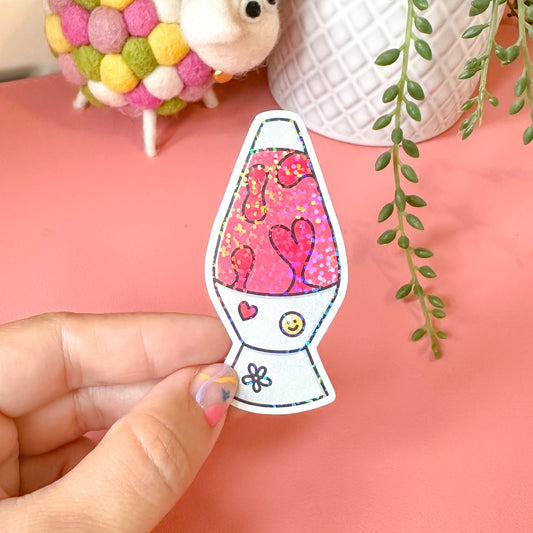 Hand holding a colorful lava lamp shaped sticker with a pink interior, surrounded by decorative items on a pink surface.