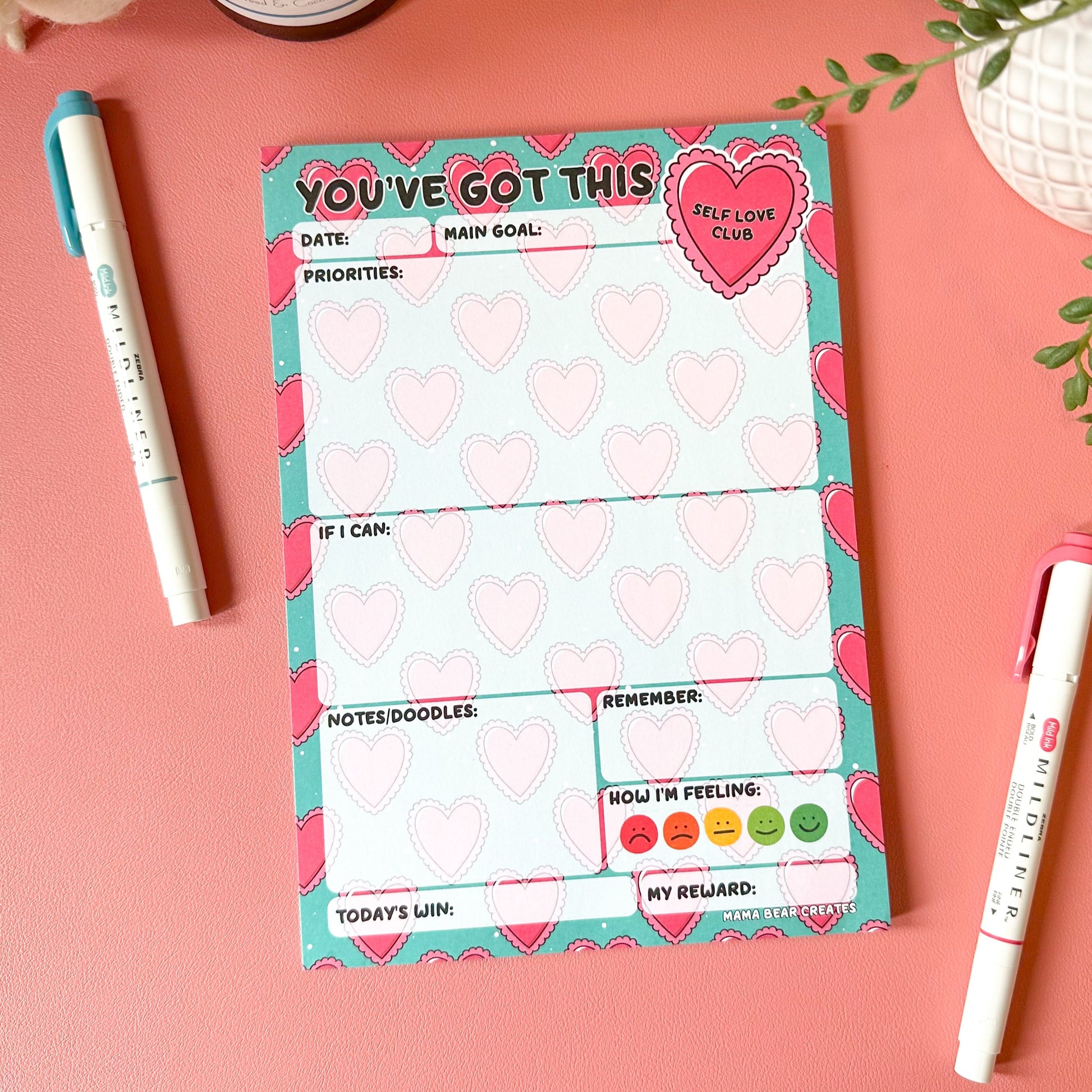 Motivational notepad with heart designs on a pink surface with pens and decor items.