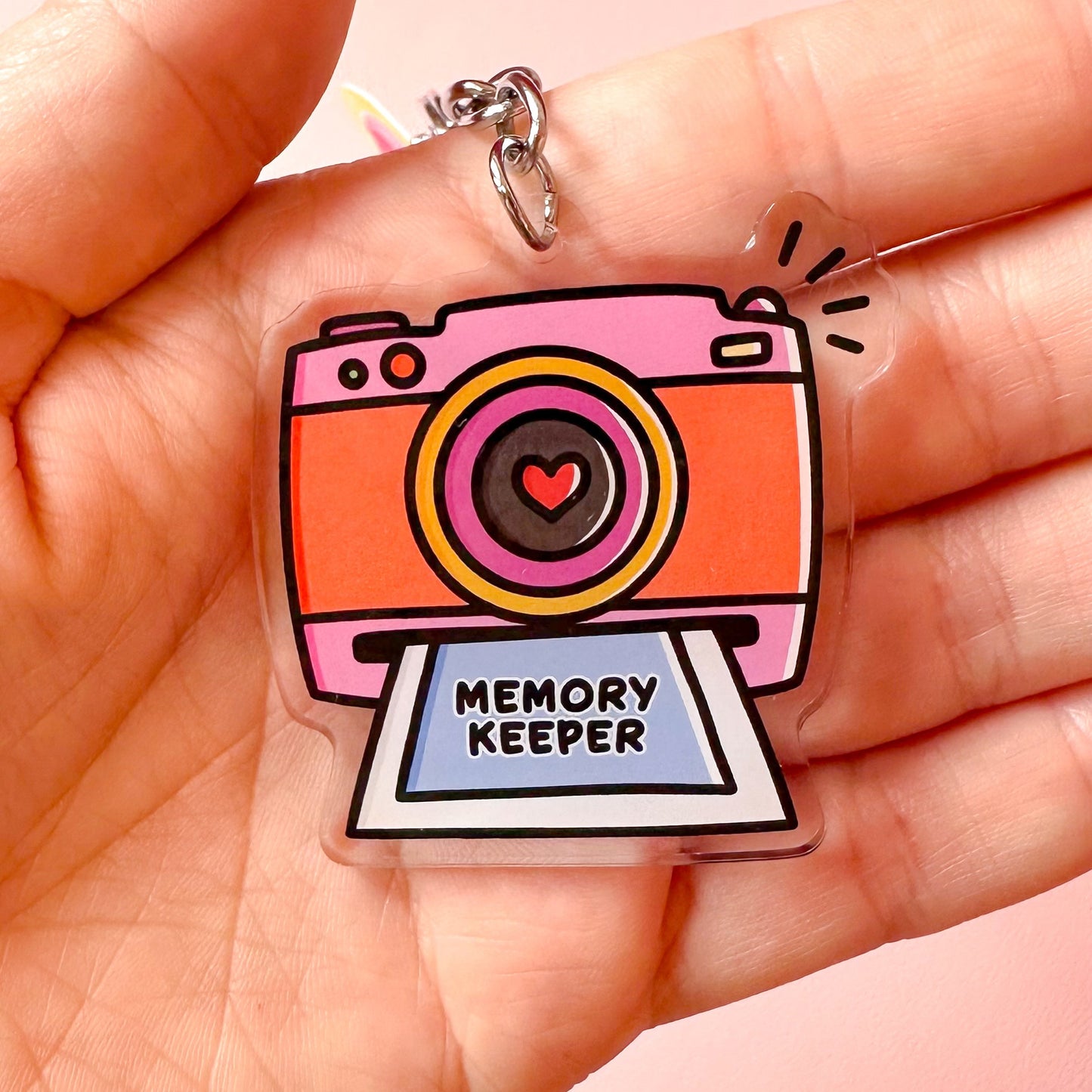 Colorful camera-shaped keychain with 'Memory Keeper' text held in a hand against a pink background
