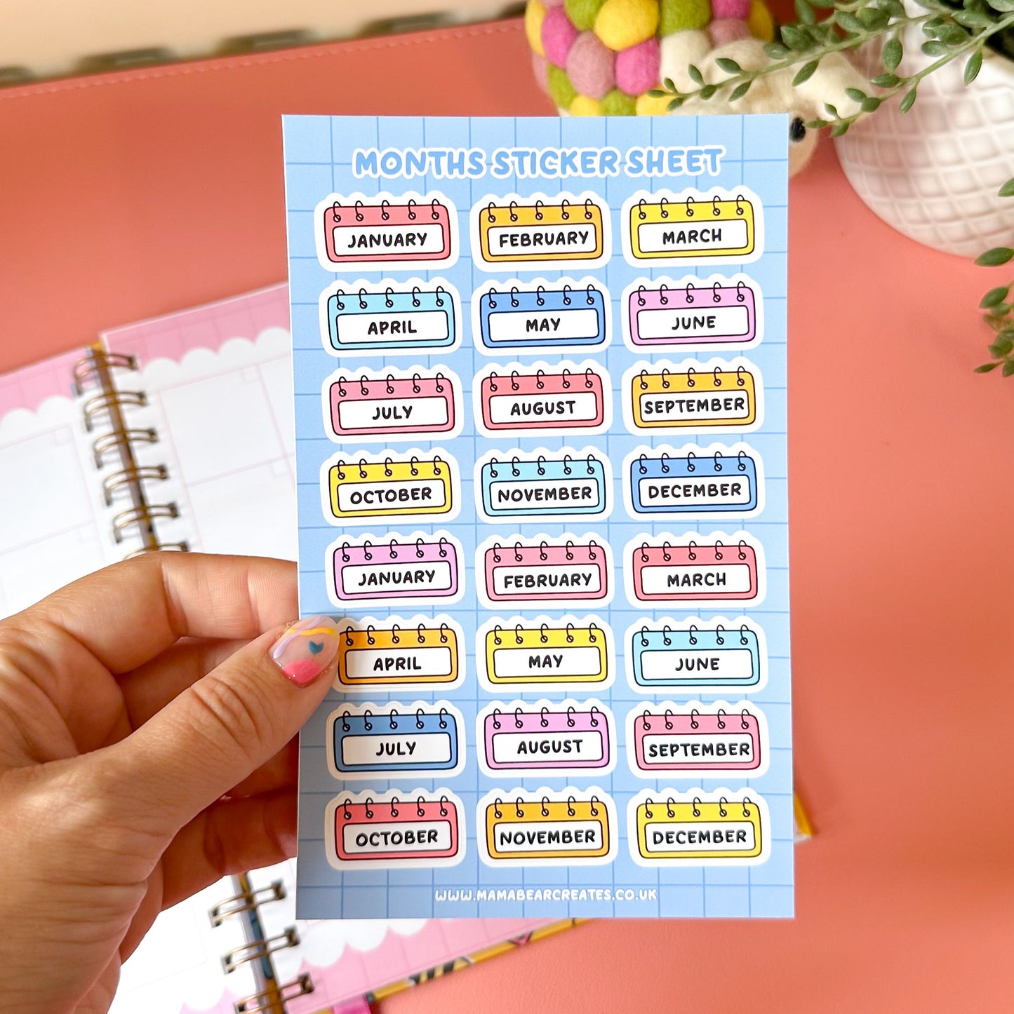 Sheet of colorful month labels held by a hand with a pink notebook in the background.