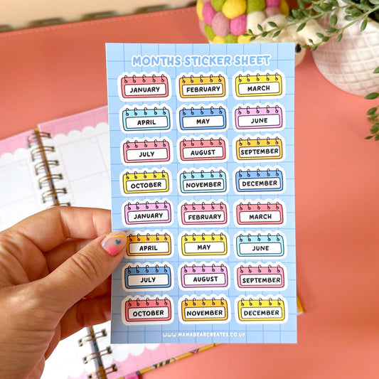 Sheet of colorful month labels held by a hand with a pink notebook in the background.