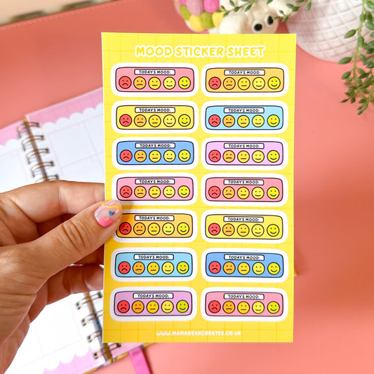 Sheet of colorful mood stickers held by a hand on a pink surface with a plant in the background.