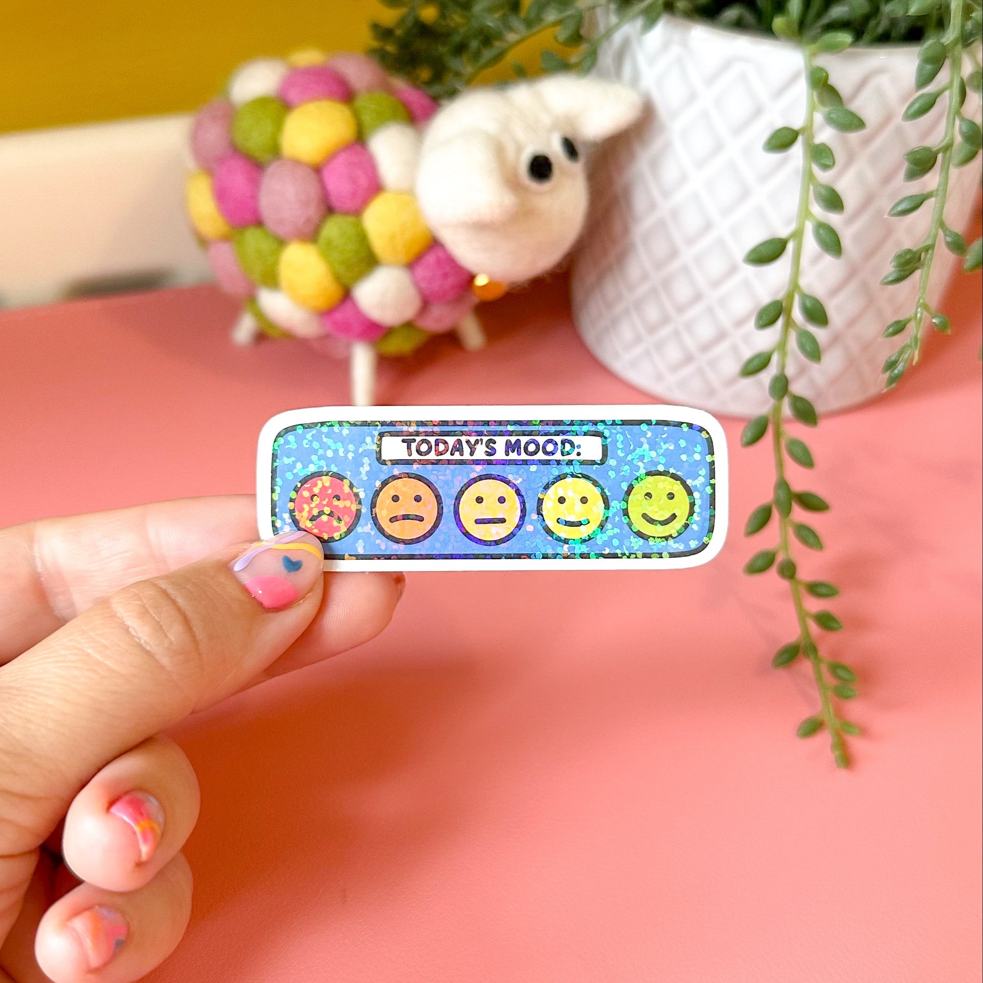 Hand holding a colorful mood sticker with emoji faces on a pink surface with a plant in the background.