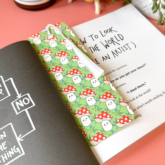 Green bookmark with mushroom pattern on a pink surface with a book and bottle in the background