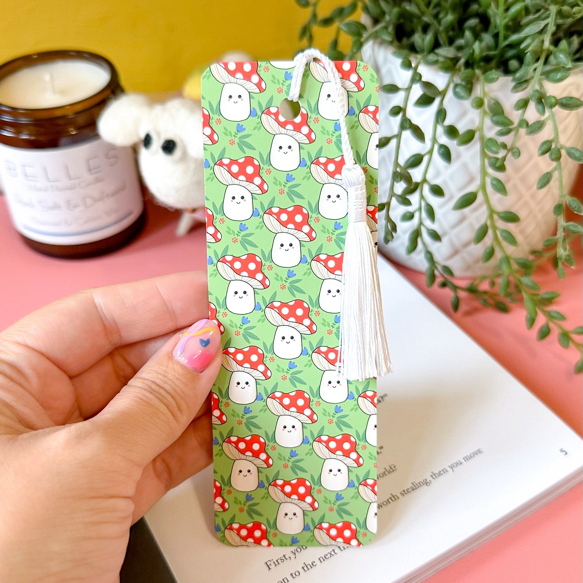 Hand holding a mushroom patterned bookmark with a yellow wall, candle, and plant in the background