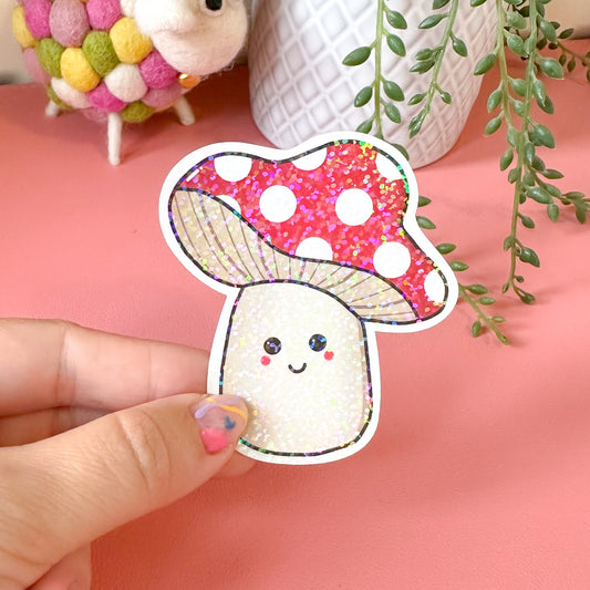 Hand holding a cute cartoon mushroom sticker with a pink polka dot cap against a pink background.