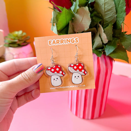 A pair of red and white mushroom-shaped earrings on an orange cardboard backing, held in a hand with a pink and orange background.