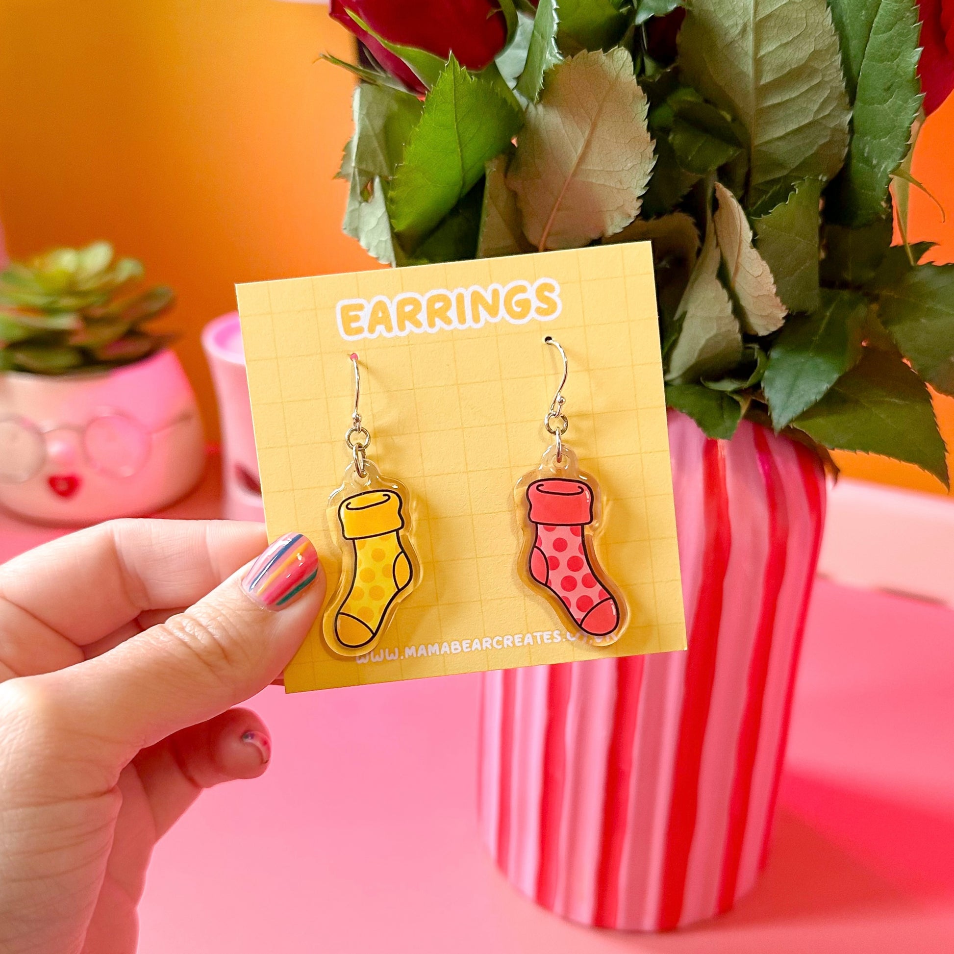 A pair of colorful acrylic earrings shaped like socks, displayed on a yellow card with the word 'EARRINGS' written on it, held by a hand with painted nails.