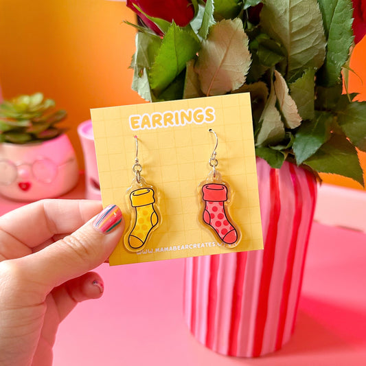 A pair of colorful acrylic earrings shaped like socks, displayed on a yellow card with the word 'EARRINGS' written on it, held by a hand with painted nails.