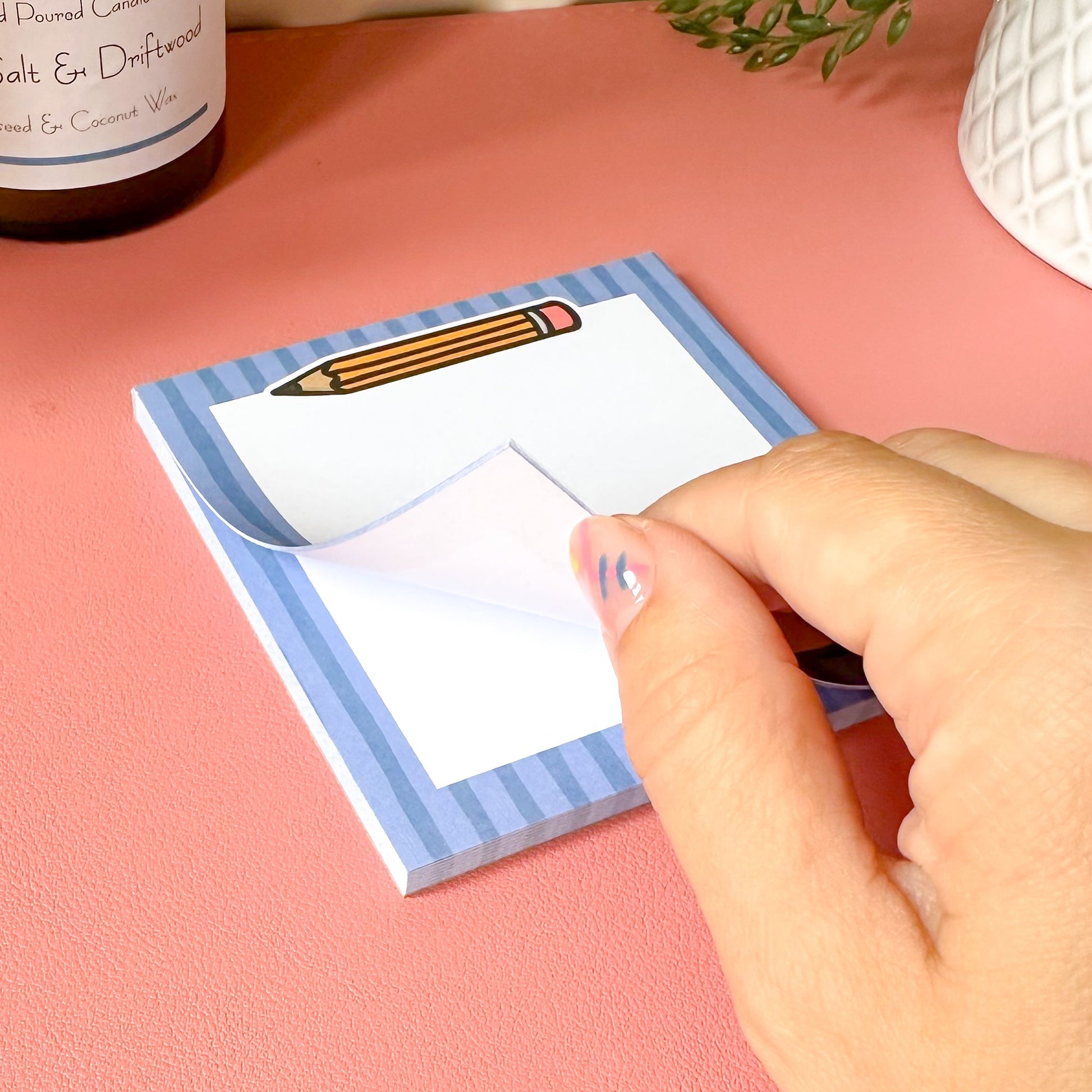 Hand holding a small notepad with a pencil design on a pink surface.