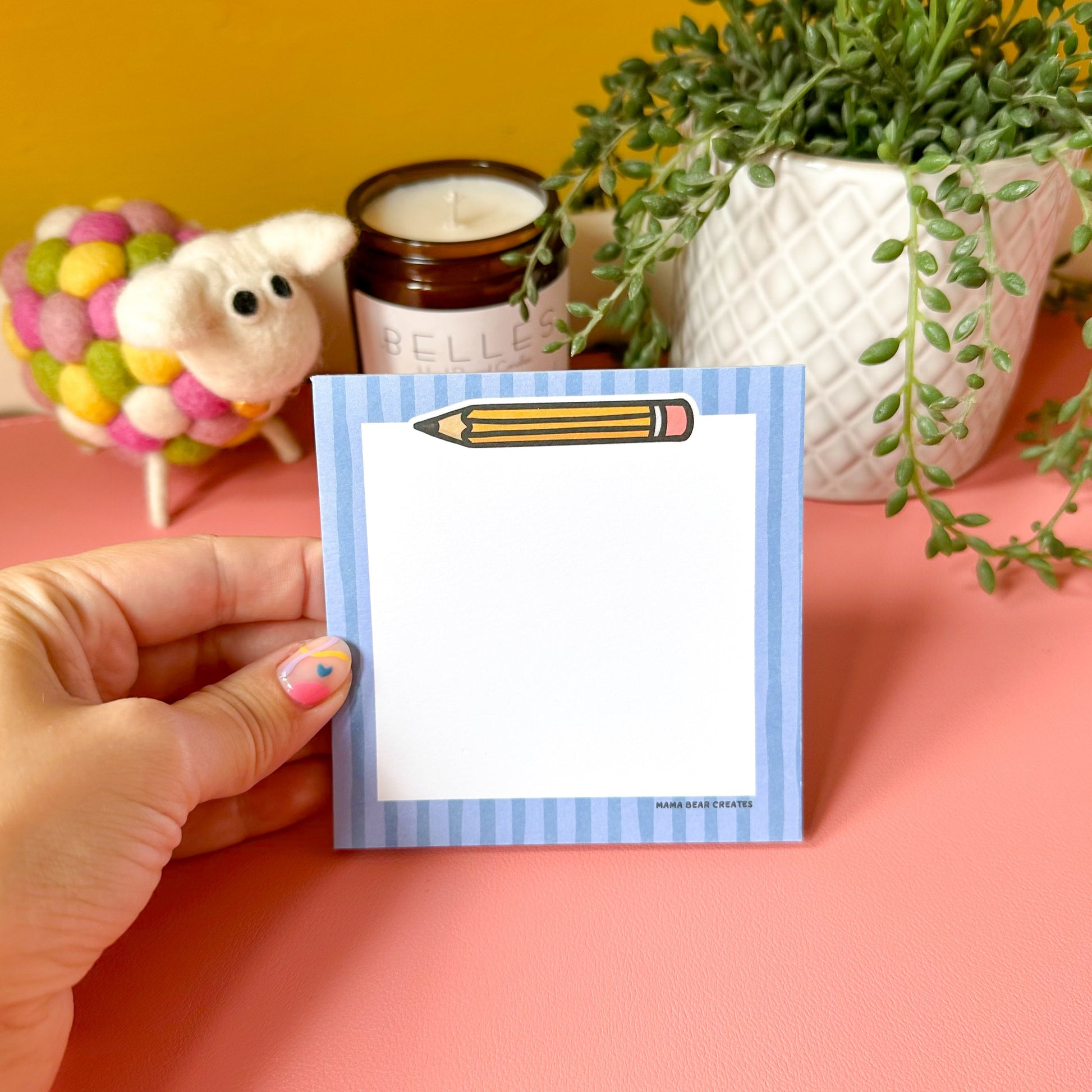Hand holding a small notepad with a pencil design on a pink surface with decorative items.