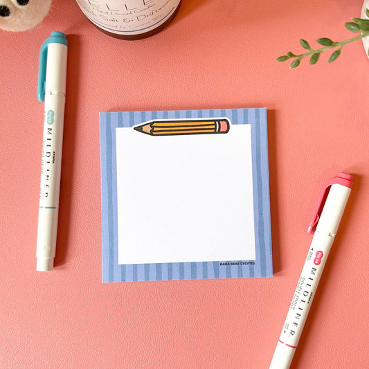 Notepad with a pencil design, pen, and candle on a pink surface