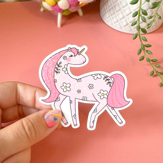 Hand holding a decorative unicorn sticker on a pink surface with a colorful ball and plant in the background.
