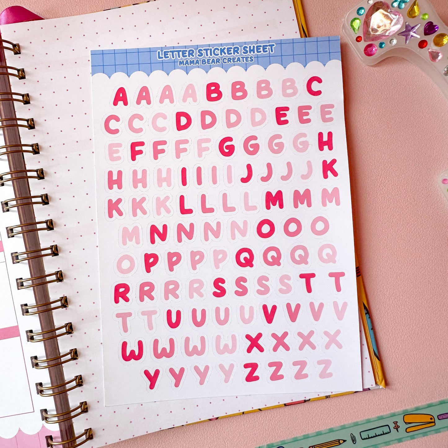 Letter sticker sheet with pink letters on a notebook with stationery items on a pink background