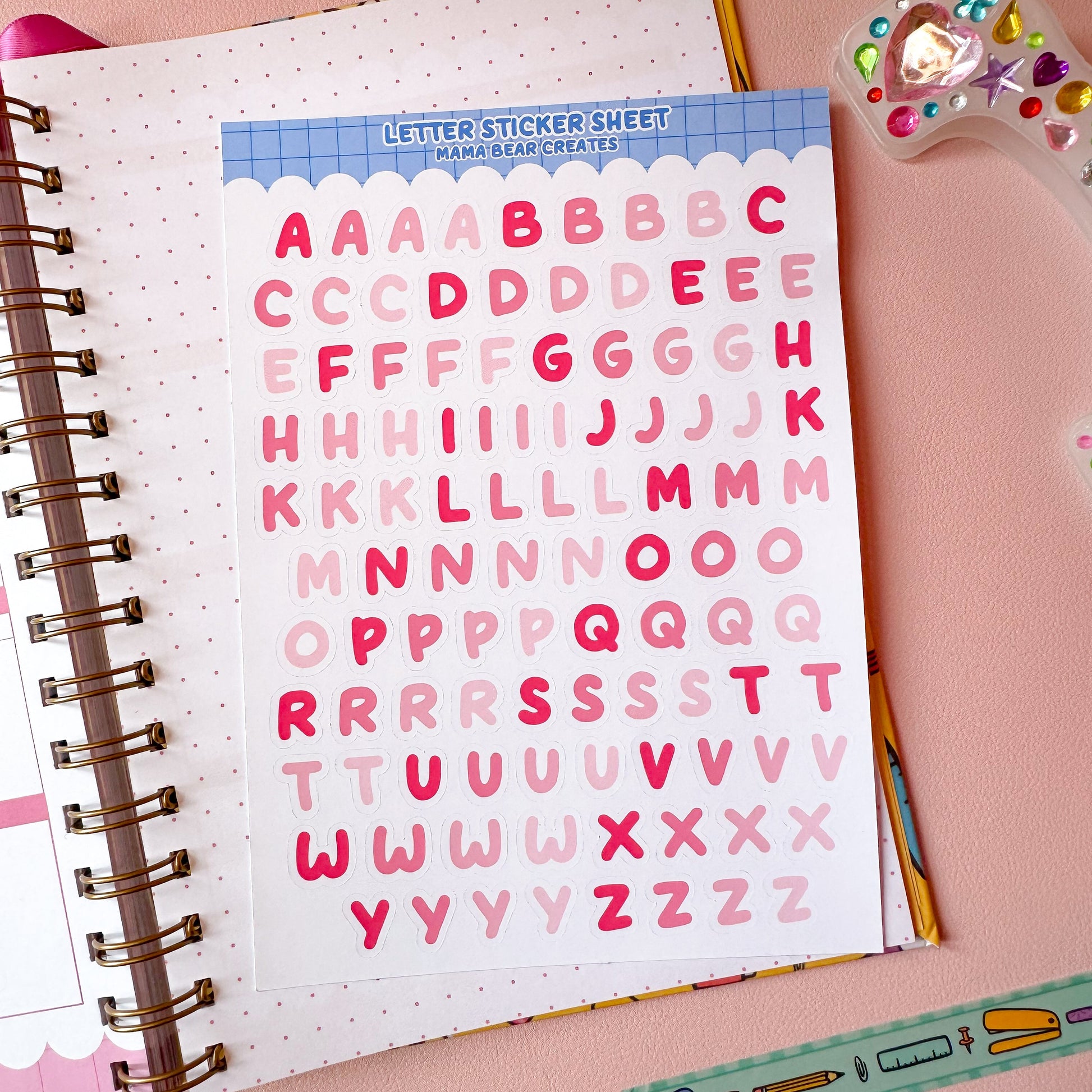 Letter sticker sheet with pink letters on a notebook with stationery items on a pink background