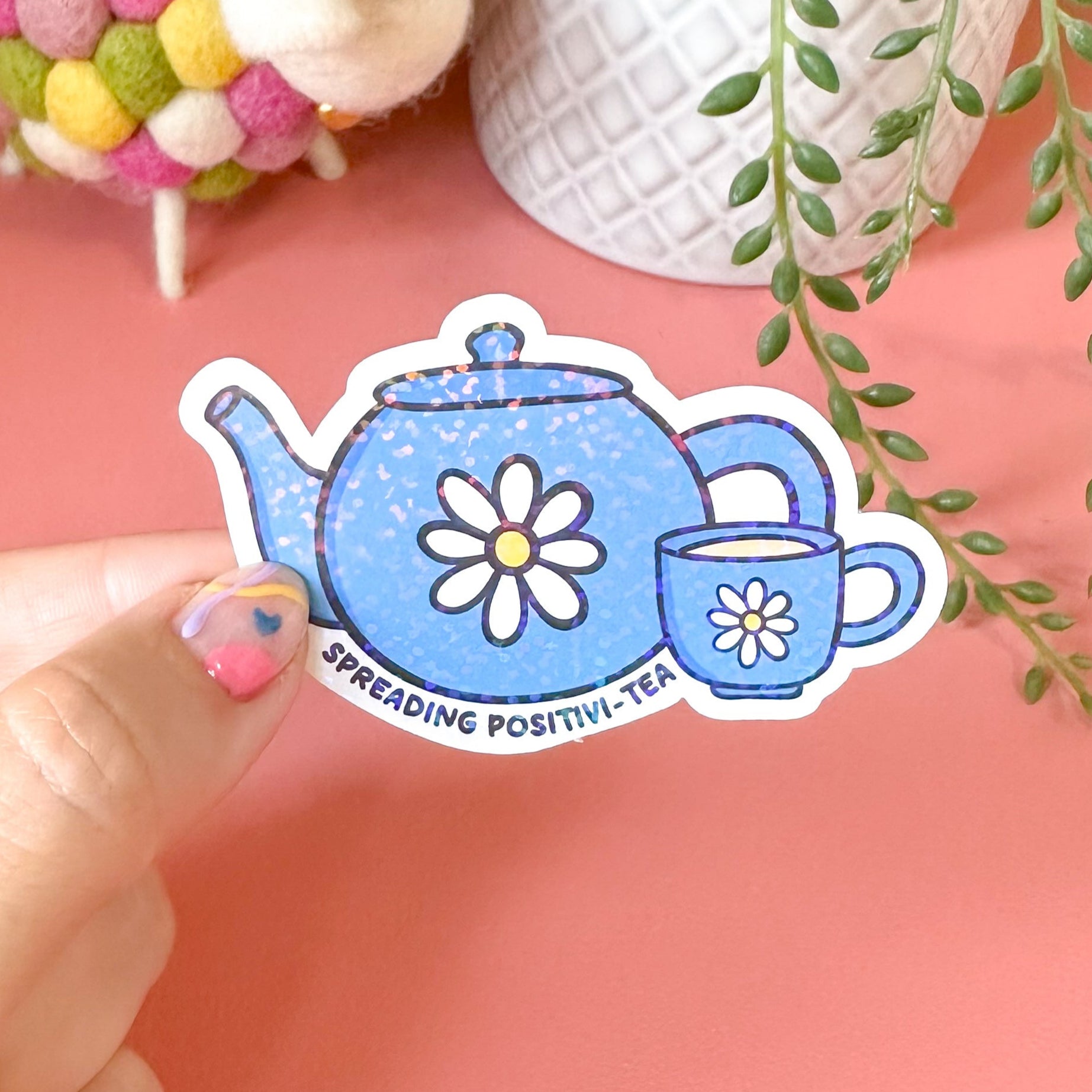 Hand holding a decorative sticker of a teapot and cup with plants and a sheep toy in the background.