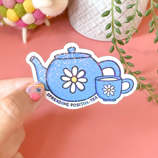 Hand holding a decorative sticker of a teapot and cup with plants and a sheep toy in the background.