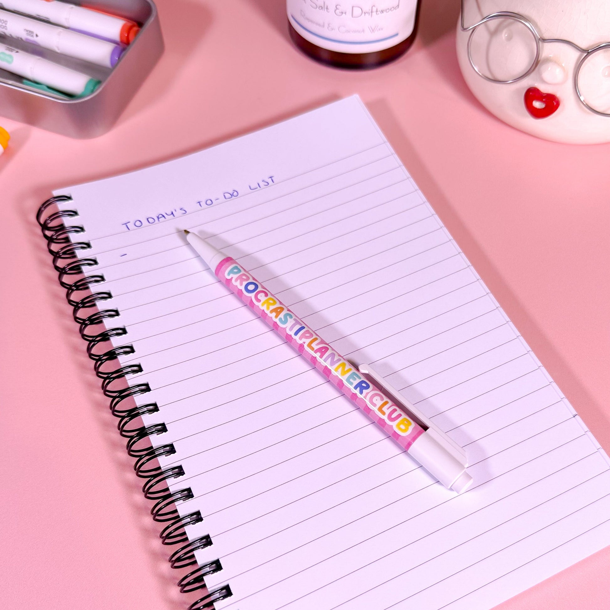 Notebook with a pen on a pink surface, surrounded by stationery items.