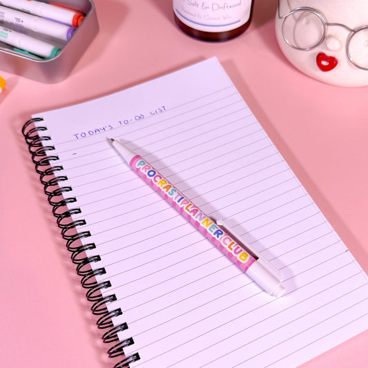 Notebook with a pen on a pink surface, surrounded by stationery items.