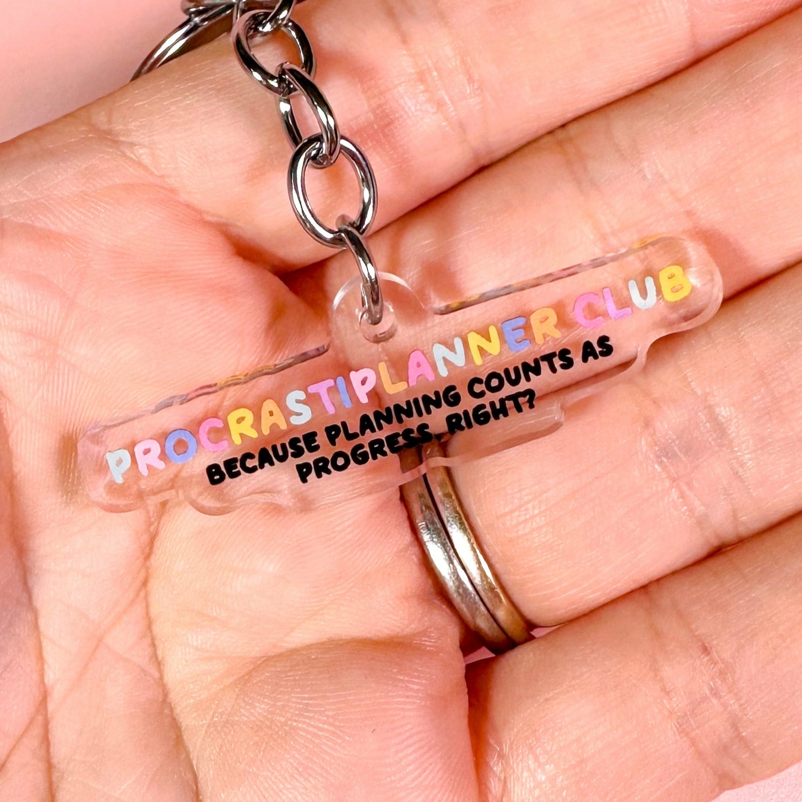 Keychain with 'Procrastiplanner Club' text held in a hand against a pink background