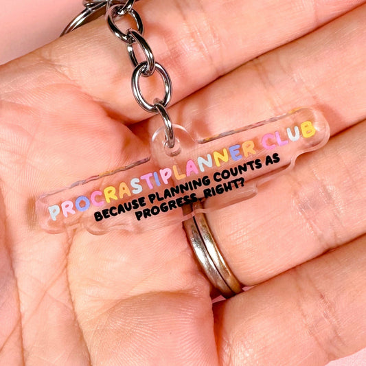 Keychain with 'Procrastiplanner Club' text held in a hand against a pink background