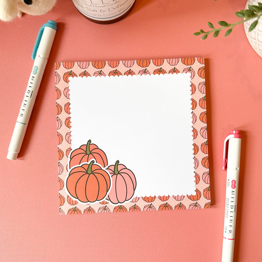 Notepad with pumpkin design on a pink surface with pens and a candle.