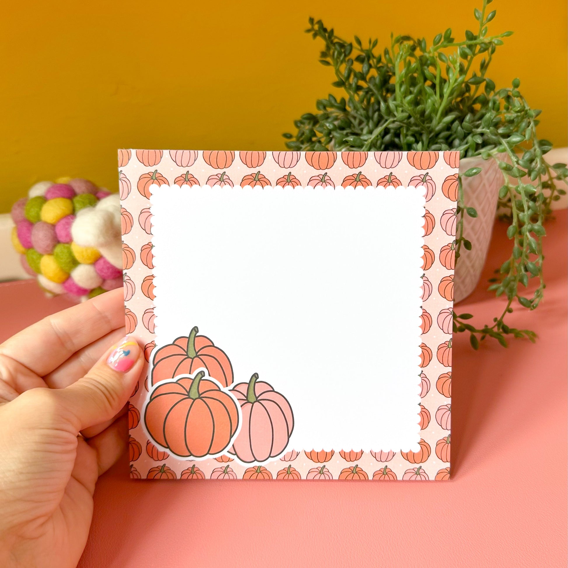 Hand holding a card with pumpkin design against a yellow wall with plants.
