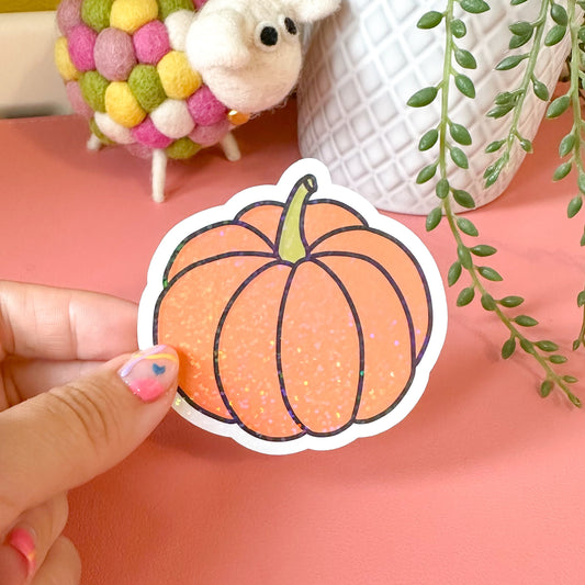Hand holding a pumpkin sticker with a pink background, toy, and plant.