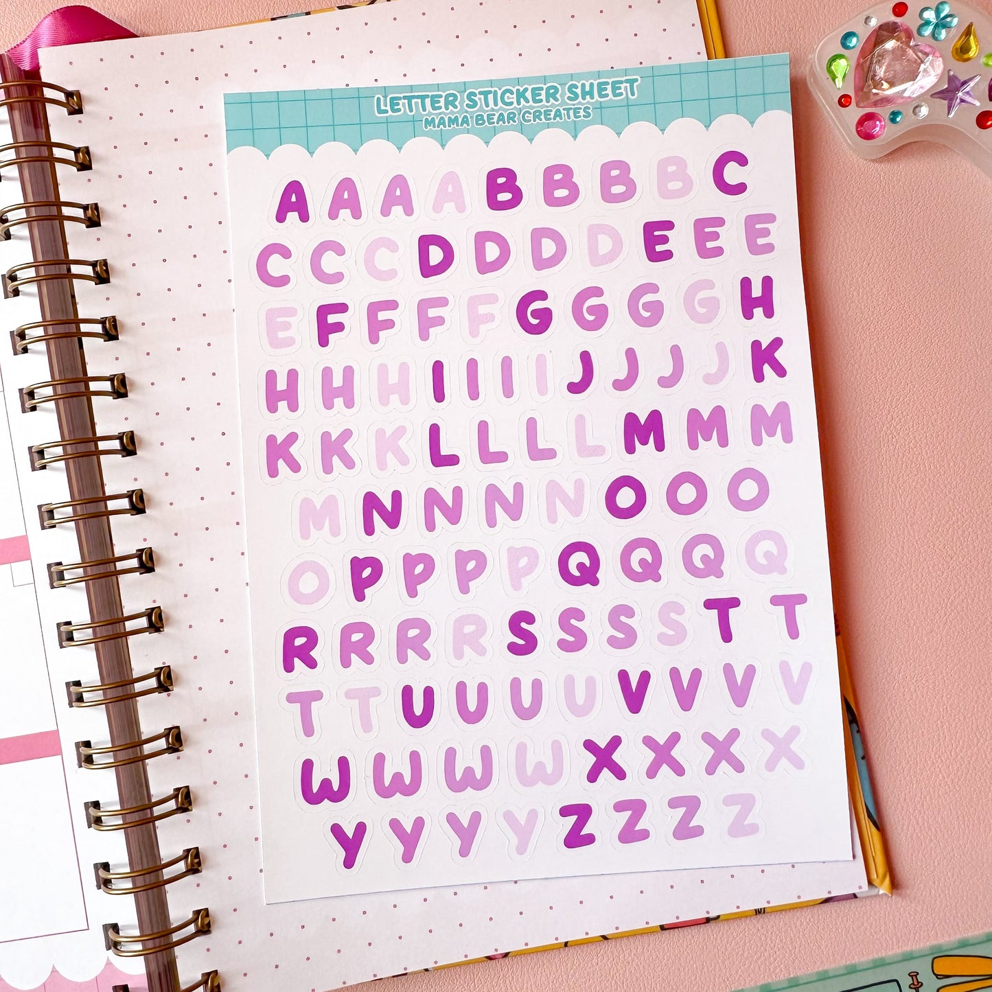 Sheet of letter stickers with a pink and purple color scheme on a pink surface with stationery items.