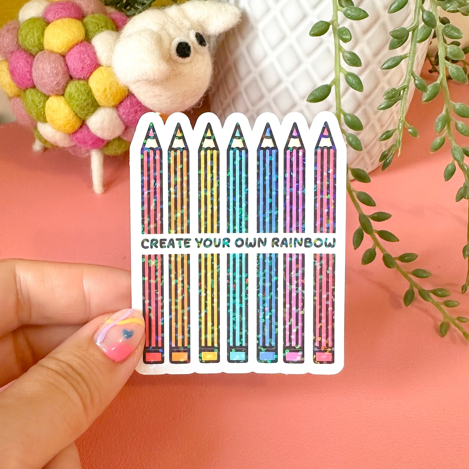 Sticker with colorful pencils and text held by a hand against a pink background with decorative elements.