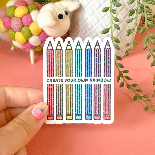 Sticker with colorful pencils and text held by a hand against a pink background with decorative elements.