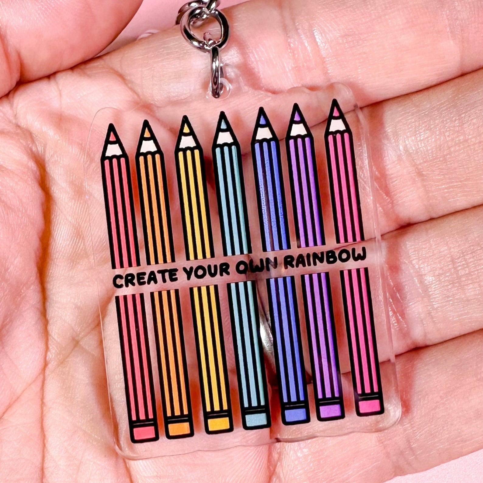 Keychain with colorful pencils and text 'Create Your Own Rainbow' held in a hand.