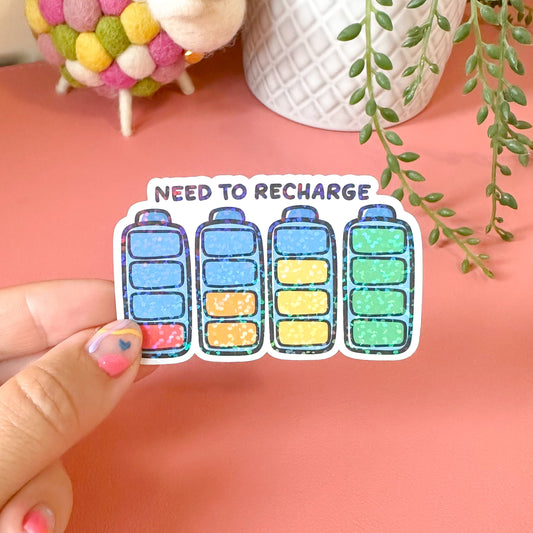 Sticker with 'NEED TO RECHARGE' text held by a hand on a pink surface with a plant in the background.