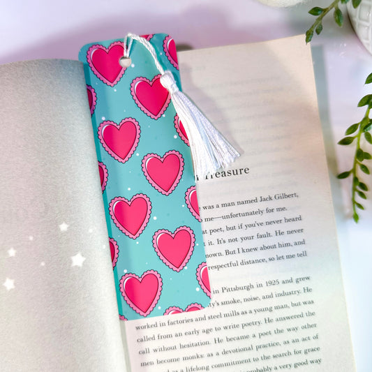 A scalloped heart-patterned bookmark with a blue background and pink hearts, attached to a book page, with a white tassel.
