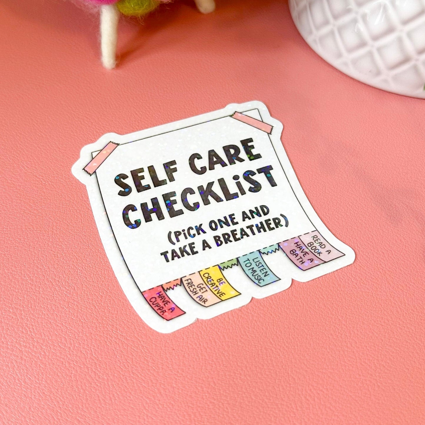 Self care checklist sticker on a pink surface with decorative items in the background