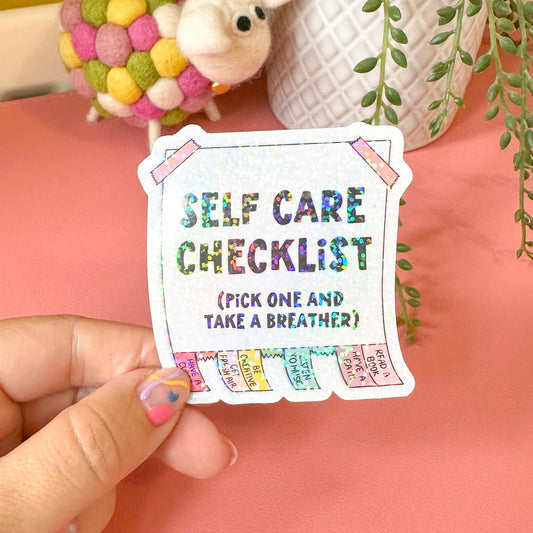 Hand holding a colorful 'Self Care Checklist' card with decorative items in the background.