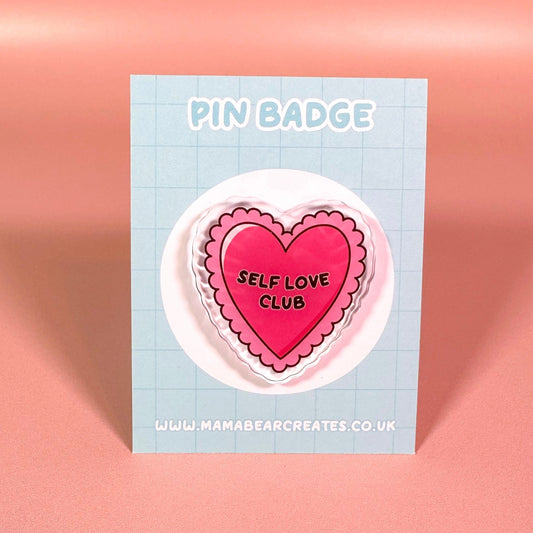 Heart-shaped pin badge with 'Self Love Club' text on a blue card against a pink background