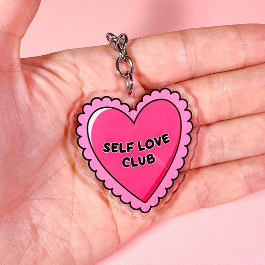 Heart-shaped 'Self Love Club' charm held in a hand against a pink background