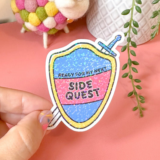 Hand holding a colorful sticker with text, small sheep toy, and plant in the background