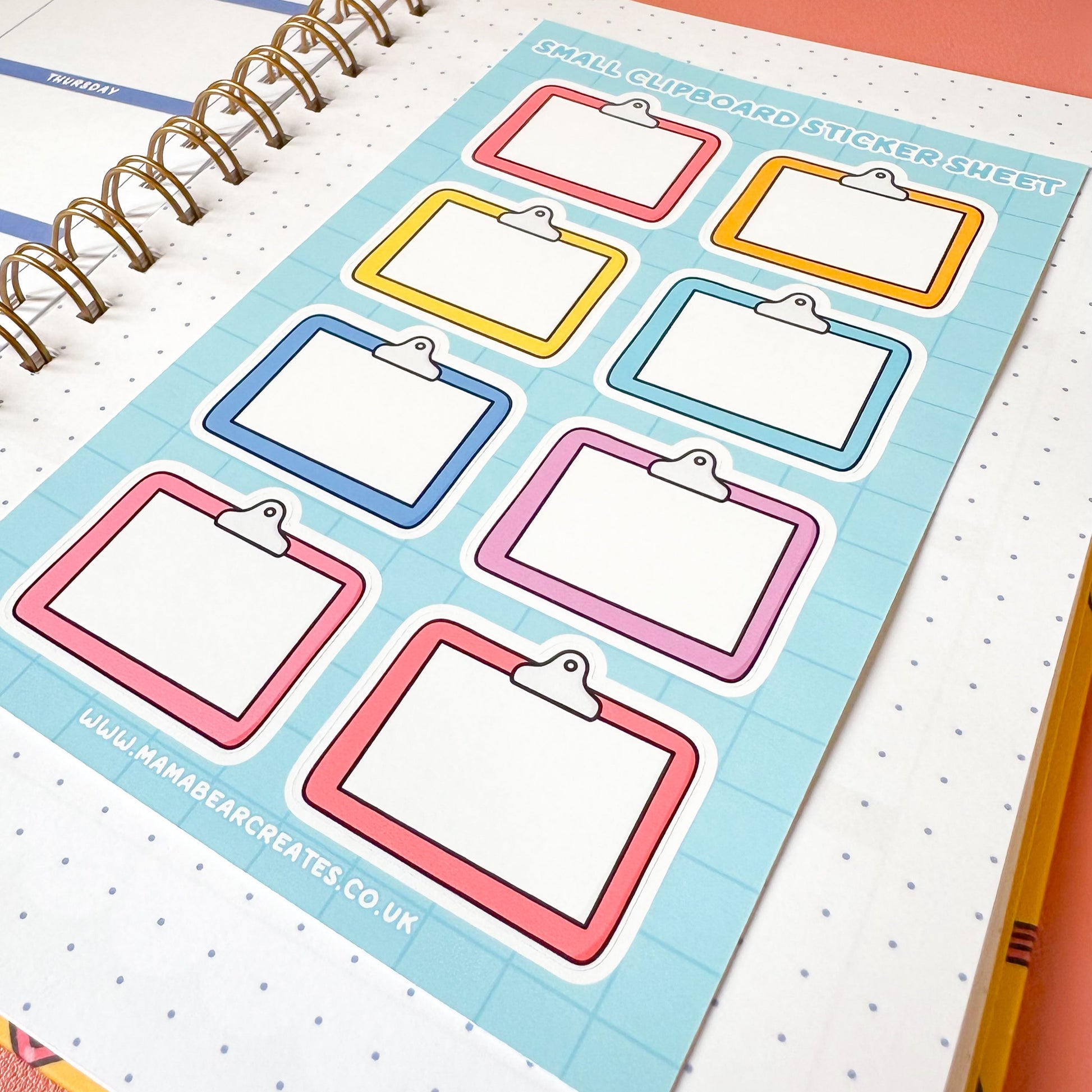 Colorful clipboard stickers on a planner page with a pen on a pink surface.
