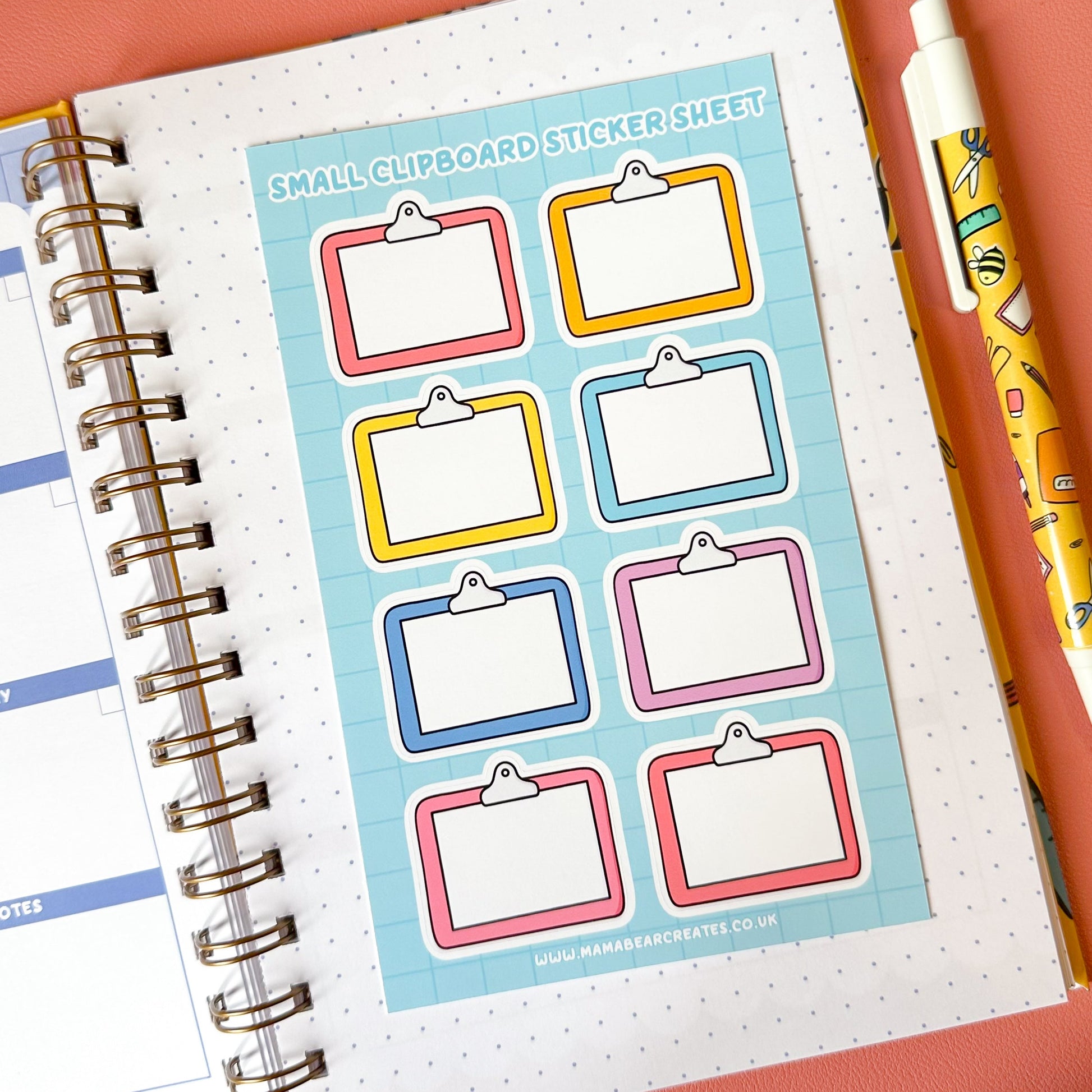 Sheet of small clip and card stickers on a planner with a pen and plant on a pink background
