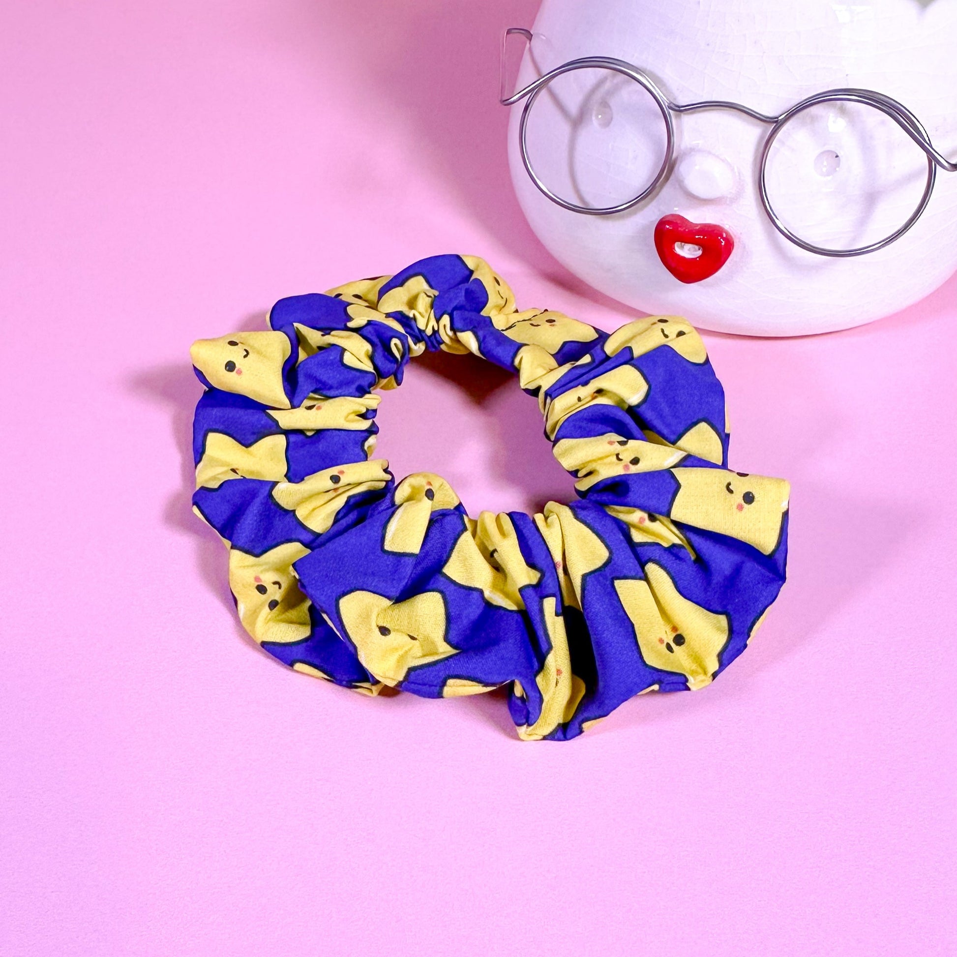 Blue and yellow scrunchie on a pink background with a plant and glasses in the background.