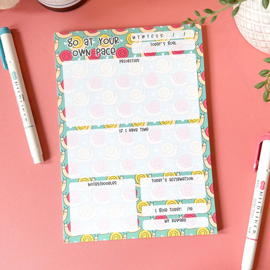 Colorful notepad with a decorative border on a pink surface with pens and a plant.