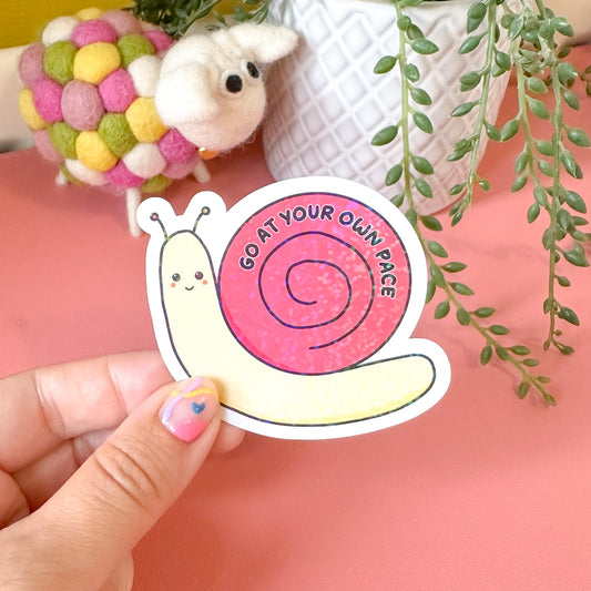 Hand holding a sticker with a snail design and text, with a pink background and green plant in the background.
