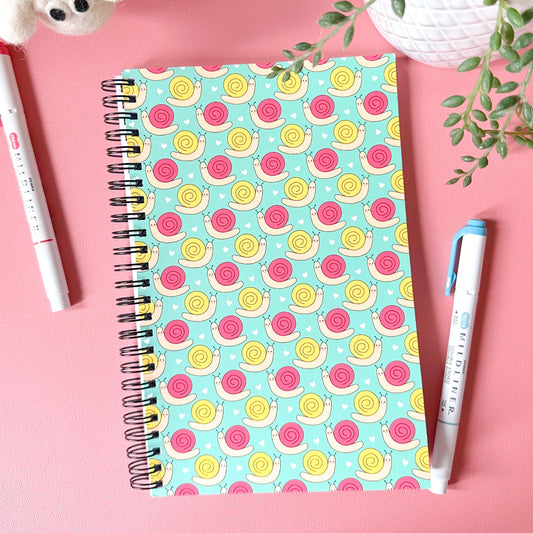 Notebook with a colorful snail pattern on a pink surface with pens and a plant.
