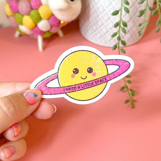 Hand holding a cute yellow planet sticker with pink orbit on a pink background