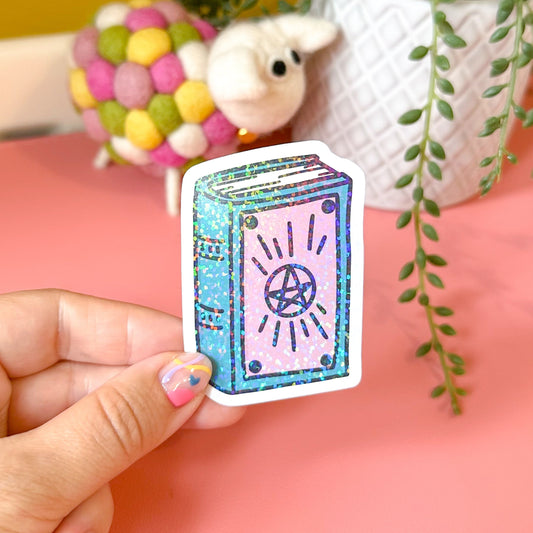 Hand holding a colorful sticker of a book with a pentacle symbol against a pink background.