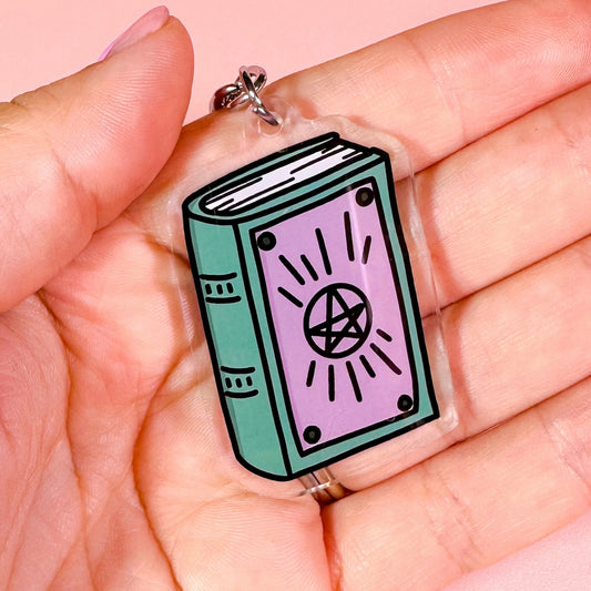 Hand holding a keychain with a book design featuring a pentagram symbol on a pink background